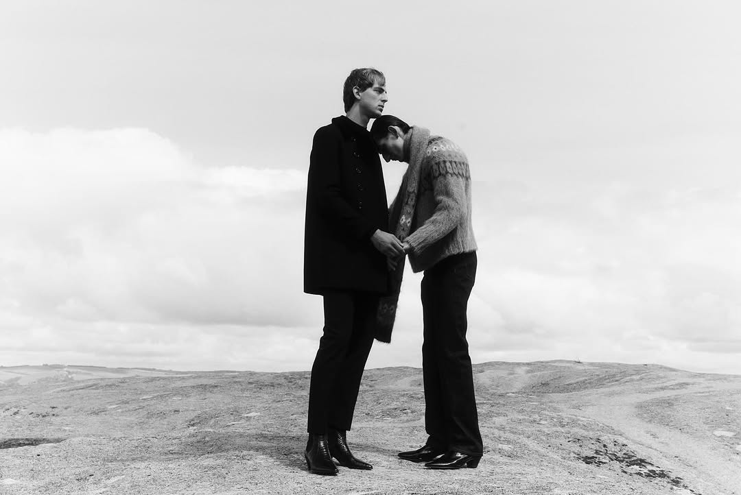 Black-and-white portrait of a couple standing on a barren landscape. One man leans his head on the other's shoulder in this outdoor scene.