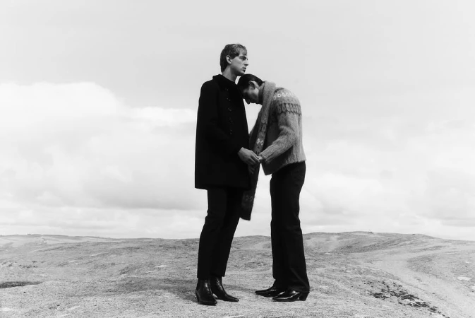 Black-and-white portrait of a couple standing on a barren landscape. One man leans his head on the other's shoulder in this outdoor scene.