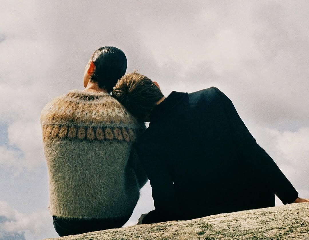 Two people viewed from behind seated closely against an outdoor sky background