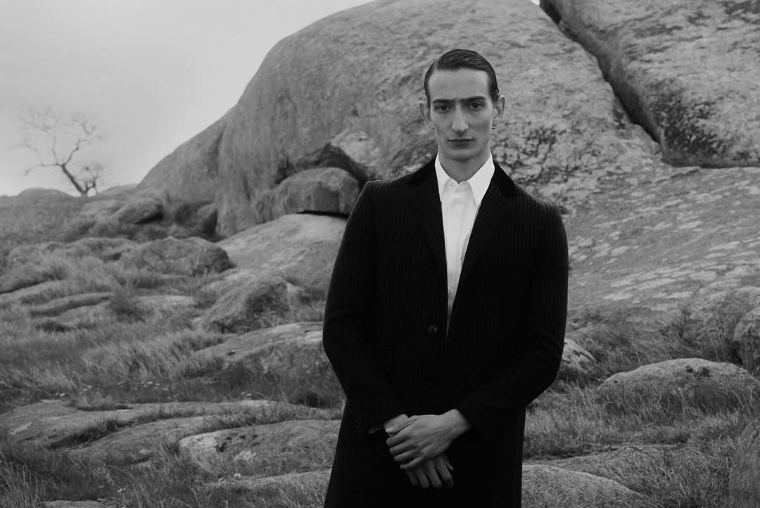 Man with slicked-back hair wearing a dark coat and white shirt against a rocky outcrop in To Be Magazine Post-War & Love editorial