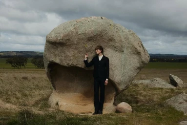 Man in a dark suit standing next to a massive rock boulder in a grassy landscape under an overcast sky.