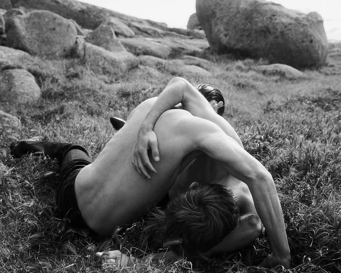 Two shirtless people embracing while lying in tall grass among large boulders outdoors in black and white.