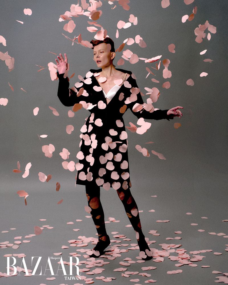 Fashion shot of a woman in black and pink polka dot dress standing amidst falling pink confetti in a studio setting, editorial.
