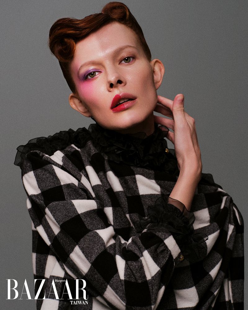 Close-up fashion portrait of a person with red hair and bold makeup, wearing a black and white checkered jacket against a gray background.