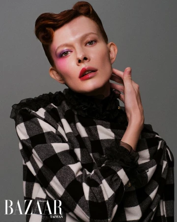 Close-up fashion portrait of a person with red hair and bold makeup, wearing a black and white checkered jacket against a gray background.