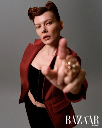 Woman in red blazer and black top strikes bold pose with retro hairstyle and statement ring in a high-fashion studio portrait.