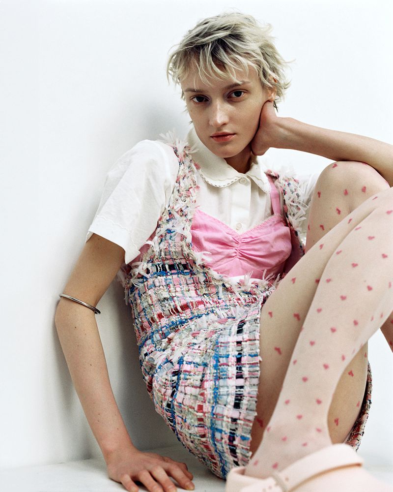 Young woman with short blonde hair poses in a colorful tweed dress, pink top, and patterned tights against a plain white backdrop.