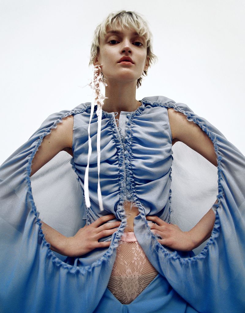 Young woman with short blonde hair poses wearing a light blue ruffled top and pants in a studio environment with a white background.