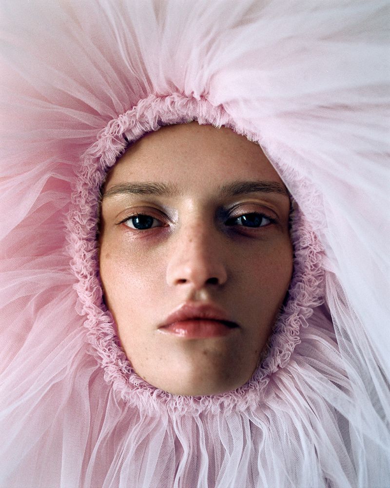 Headshot of a person with fair skin framed by a large, fluffy pink tulle headpiece. Focus is on the face, lips, and eyes.