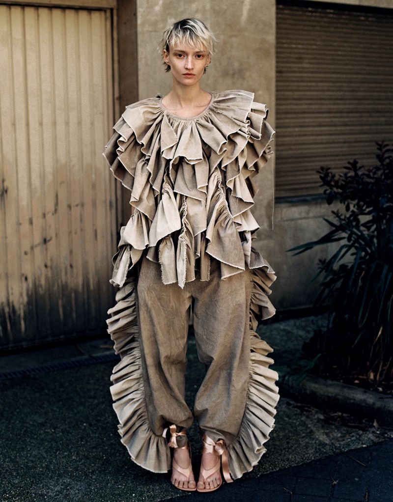 Fashion editorial shot of a woman with short blonde hair wearing a ruffled beige top and pants, standing outdoors in front of a building.