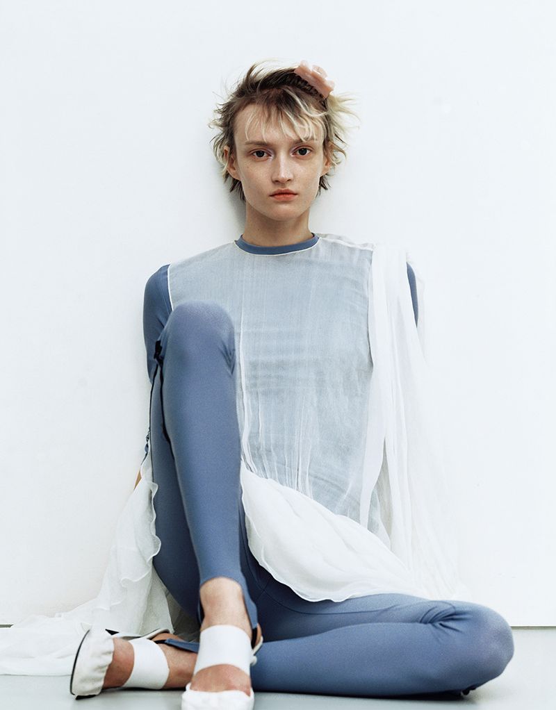 A slender woman with short blonde hair is sitting on the floor wearing a blue bodysuit and a sheer white top against a white background.