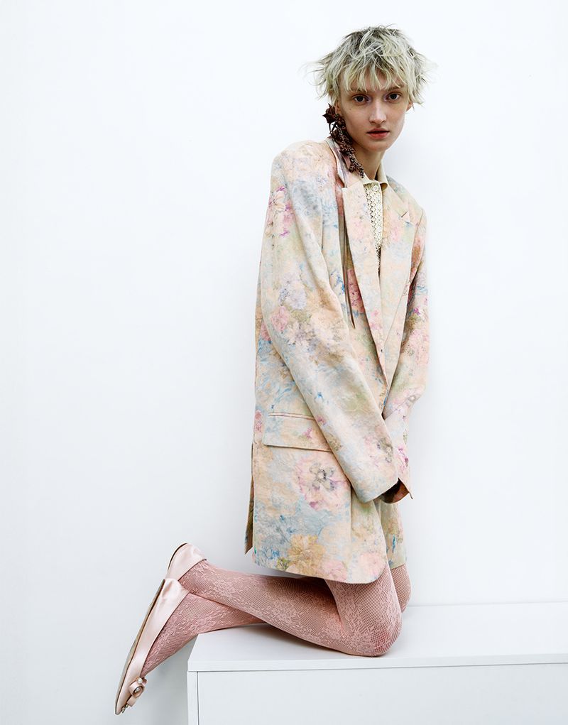 Fashion shot of a young adult woman with short blonde hair kneeling on a white surface, wearing a floral blazer and pink tights indoors.