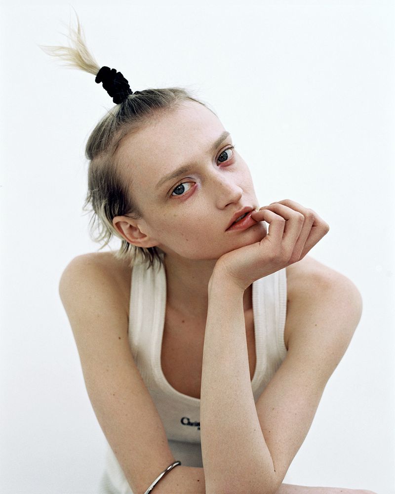 Close-up portrait of a young adult woman with blonde hair tied back, wearing a white tank top, set against a bright, neutral background.