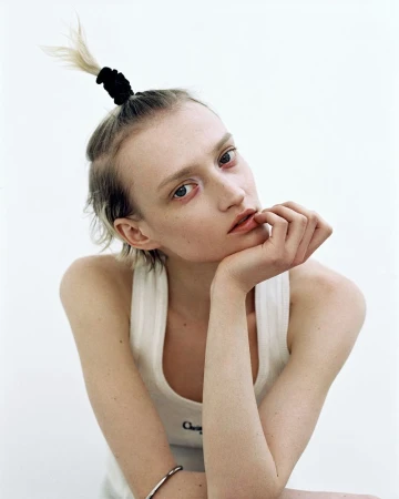 Close-up portrait of a young adult woman with blonde hair tied back, wearing a white tank top, set against a bright, neutral background.