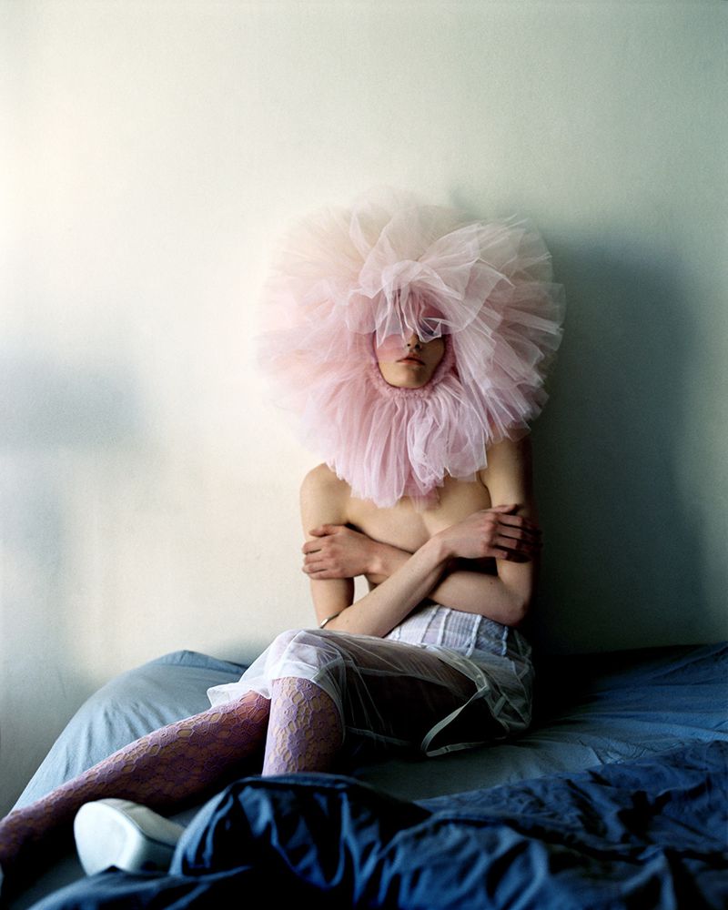 Fashion shot of person with large pink tulle collar seated on blue bed with white wall in background, creative fashion, indoor studio.