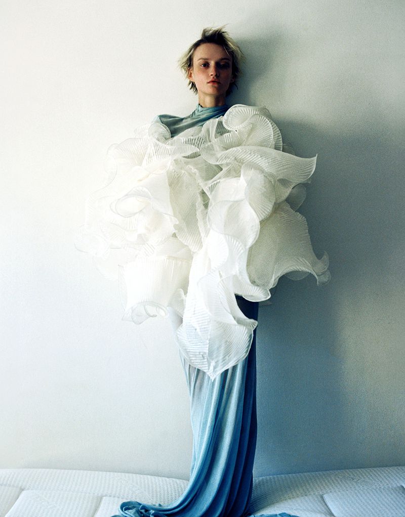 A fashion shot features a model in a blue dress with a large white ruffled garment, posing against a light wall, with a soft, artistic feel.