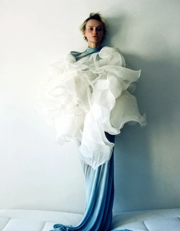 A fashion shot features a model in a blue dress with a large white ruffled garment, posing against a light wall, with a soft, artistic feel.