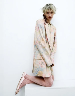 Model with short blonde hair kneeling in an oversized pastel floral blazer and pink tights against a white background.