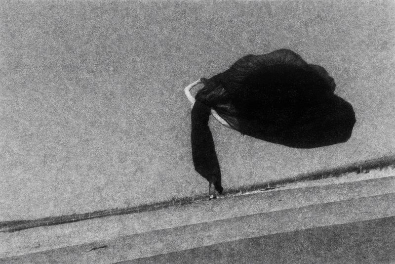 Black and white fine art photograph of a dark object, possibly a bag, placed on concrete steps in an outdoor setting under natural light.