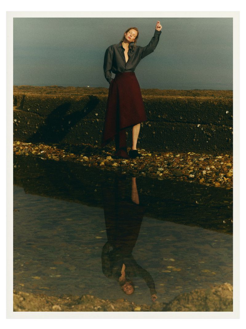 A full shot captures a woman in a gray long-sleeve shirt and maroon skirt standing on a rocky surface near a body of water with a reflection.