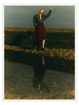 A full shot captures a woman in a gray long-sleeve shirt and maroon skirt standing on a rocky surface near a body of water with a reflection.