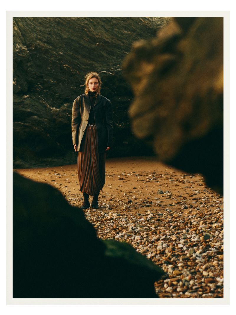 A stylish woman in a blazer and pleated skirt stands on a rocky beach with large rock formations, creating a fashion editorial scene.