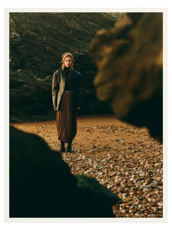 A stylish woman in a blazer and pleated skirt stands on a rocky beach with large rock formations, creating a fashion editorial scene.