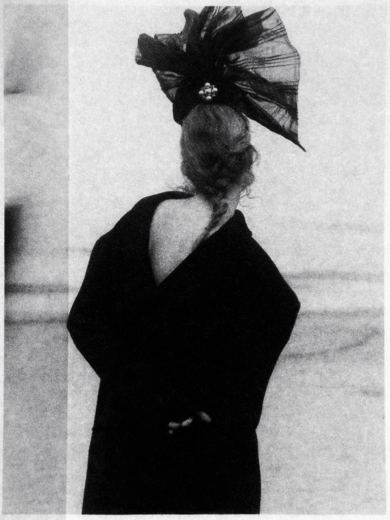 Back view of a woman with an elaborate black headpiece and elegant dark coat against a neutral background, creating a classic, high-fashion aesthetic.