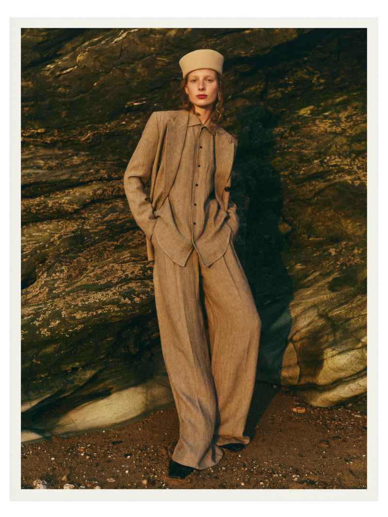 Demy de Vries standing against a rugged rocky cliff wearing a tailored beige oversized suit and structured matching hat.
