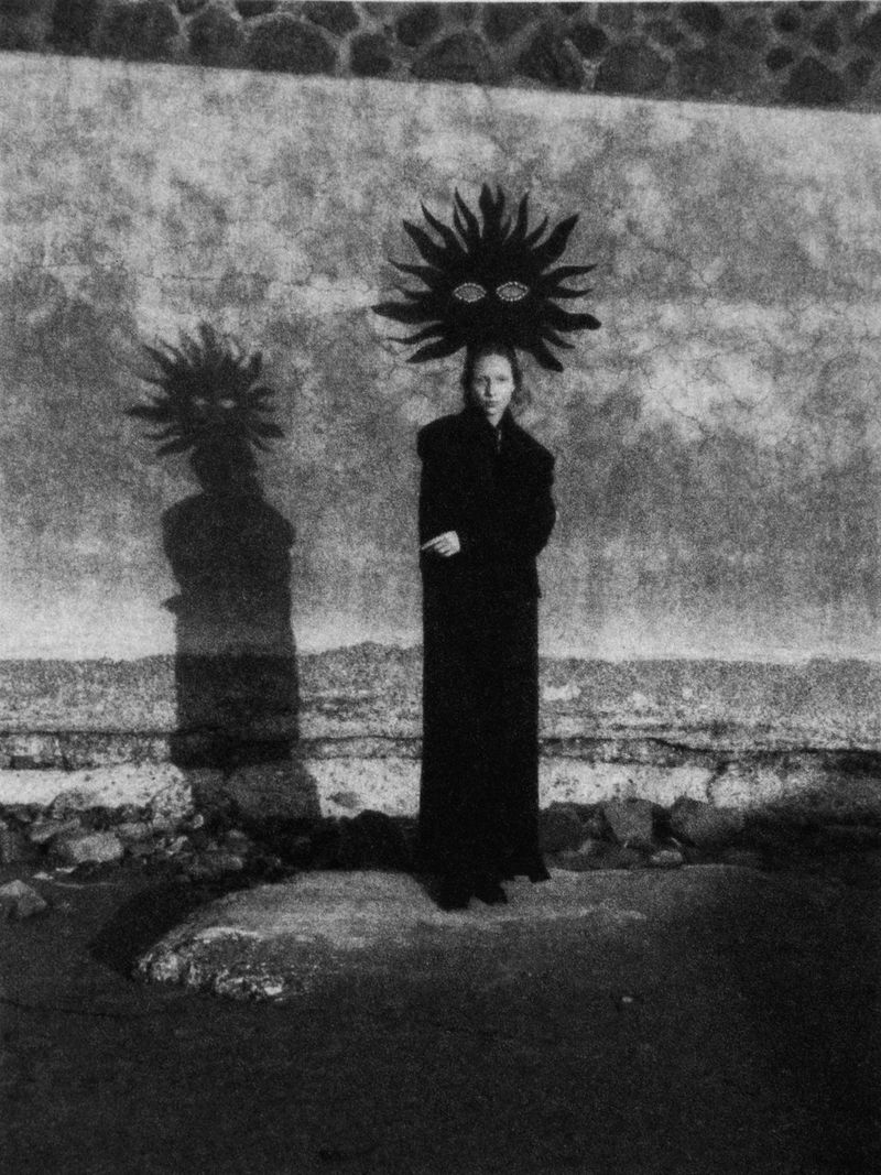 Monochrome portrait of a person in dark clothing standing in front of a textured wall, wearing an elaborate headpiece with eye details.