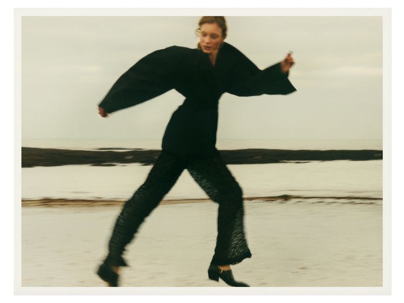 A woman in a black outfit runs on a beach with ocean water and a cloudy sky in the background in a fashion editorial style photograph.
