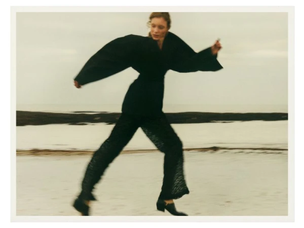 A woman in a black outfit runs on a beach with ocean water and a cloudy sky in the background in a fashion editorial style photograph.