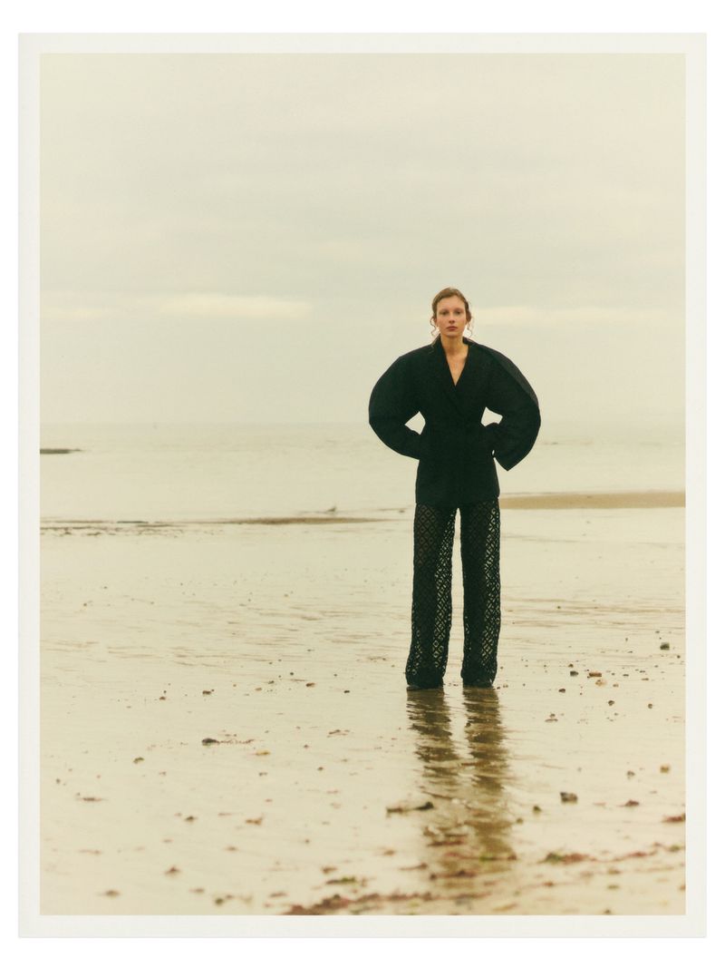 Woman in black outfit stands on sandy beach with overcast sky, her hands on her hips in a fashion editorial style portrait.