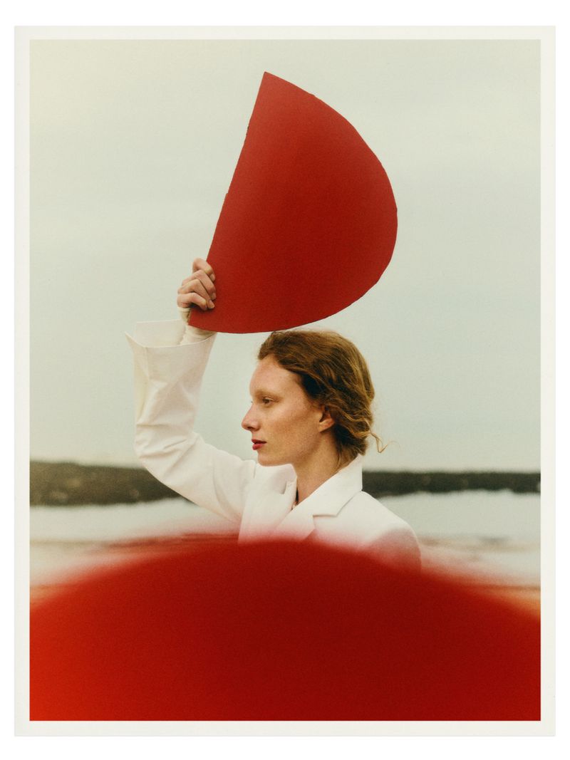 Profile of a woman in a white jacket holding a large red semi-circle against an overcast sky. A blurred semi-circle is visible at the bottom.