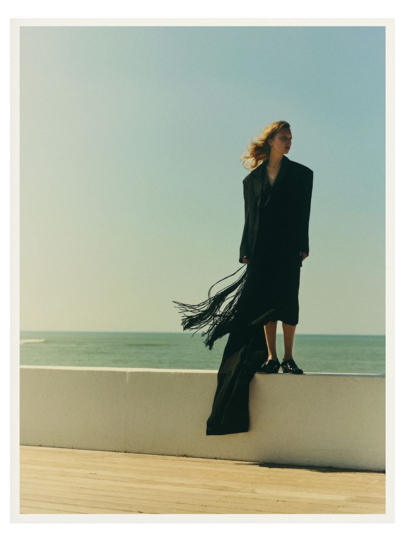 Woman in a long black coat with fringe stands on a white wall against a sea and sky backdrop in a fashion editorial style shoot.