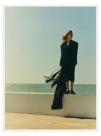 Woman in a long black coat with fringe stands on a white wall against a sea and sky backdrop in a fashion editorial style shoot.