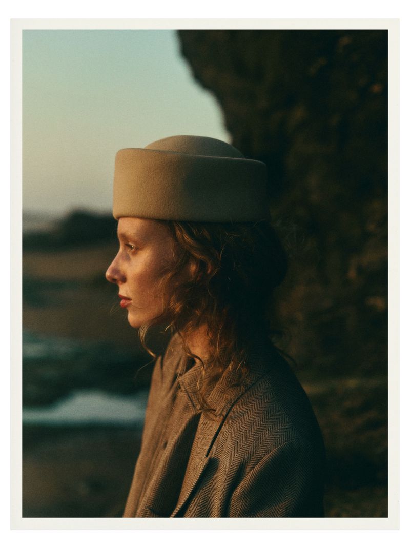 Woman in retro fashion poses outdoors with a beige hat and brown coat, featuring a close-up shot against a natural rock formation backdrop.