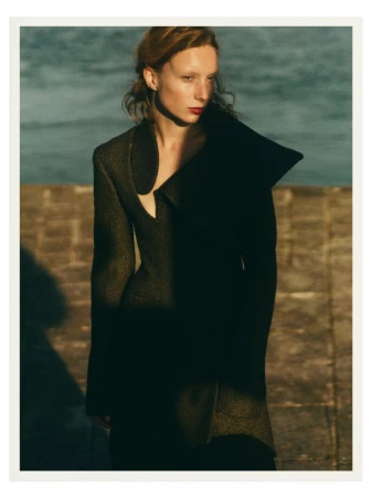 Adult woman in a gray textured jacket with dramatic lapels stands in dappled sunlight near a stone wall and body of water, outdoor fashion.