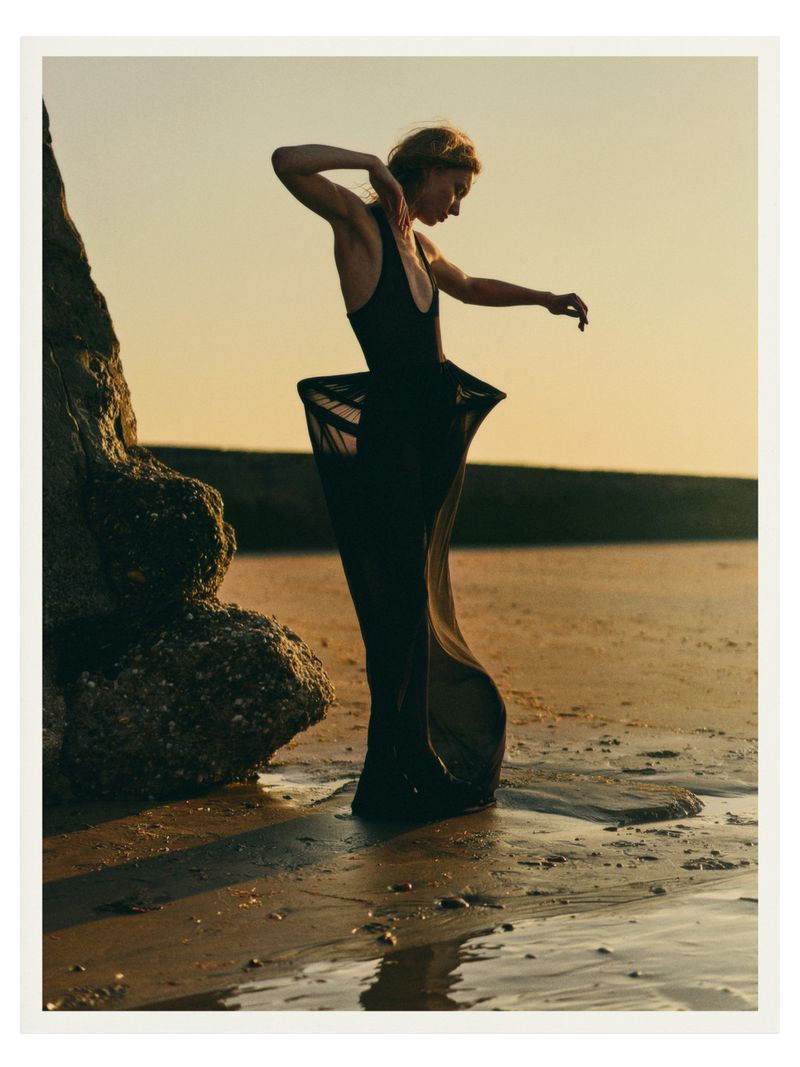 An adult female model in a black dress poses on a sandy beach next to a rock formation at sunset, lit by soft golden hour light.