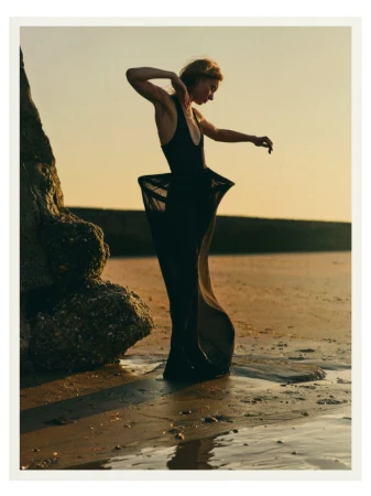 An adult female model in a black dress poses on a sandy beach next to a rock formation at sunset, lit by soft golden hour light.