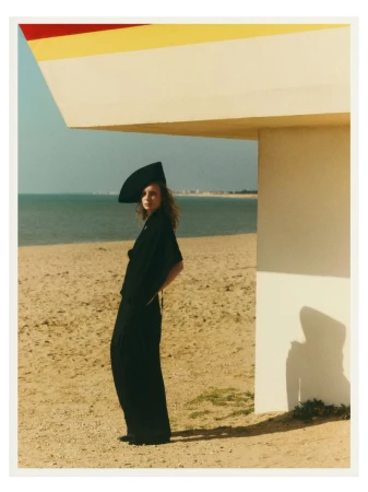 Model wearing an all-black outfit and a sculptural black headpiece standing on a sandy beach next to a pale architectural structure.