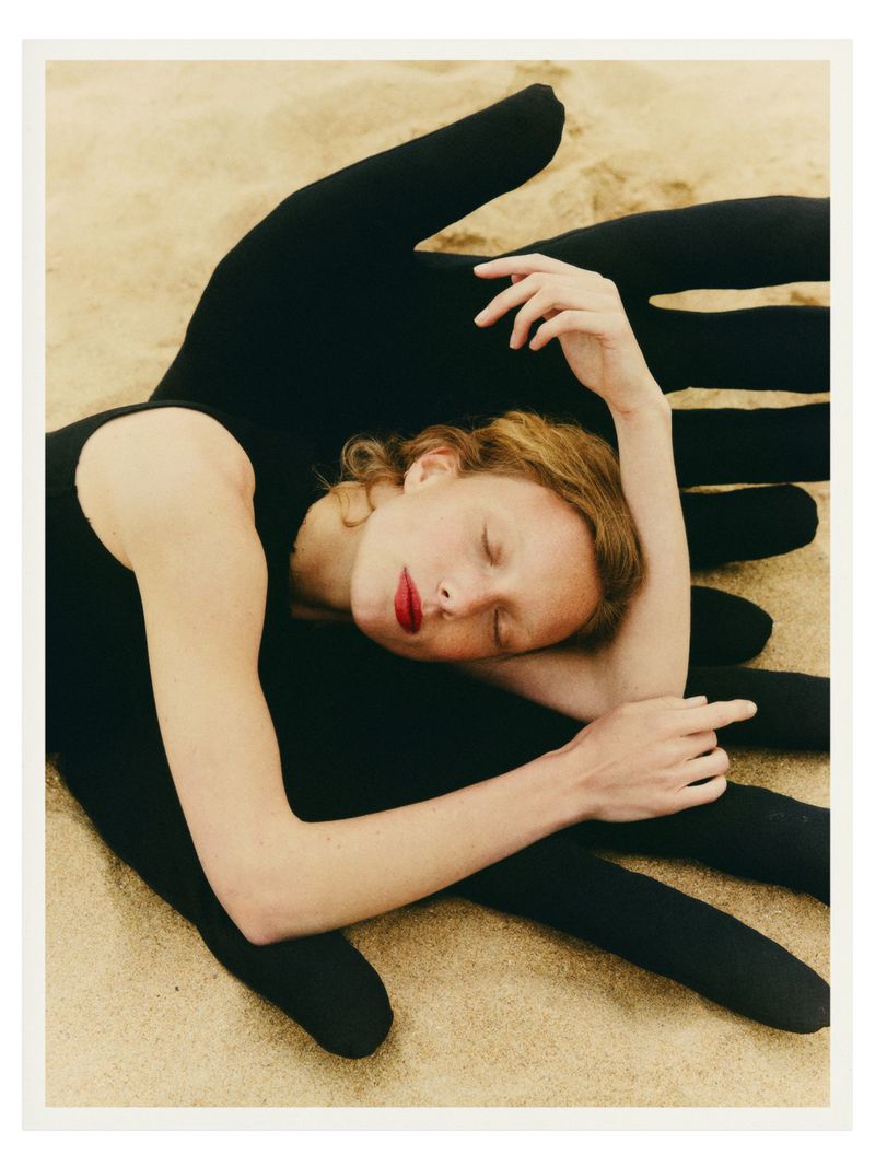 A fair-skinned woman with red lipstick rests on a black hand sculpture against a tan sand background in a studio photography setting.