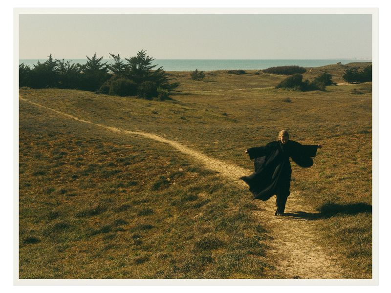 Person in black robe walks a sandy path through grassy dunes toward a distant ocean under a bright sky in a sunny outdoor setting.