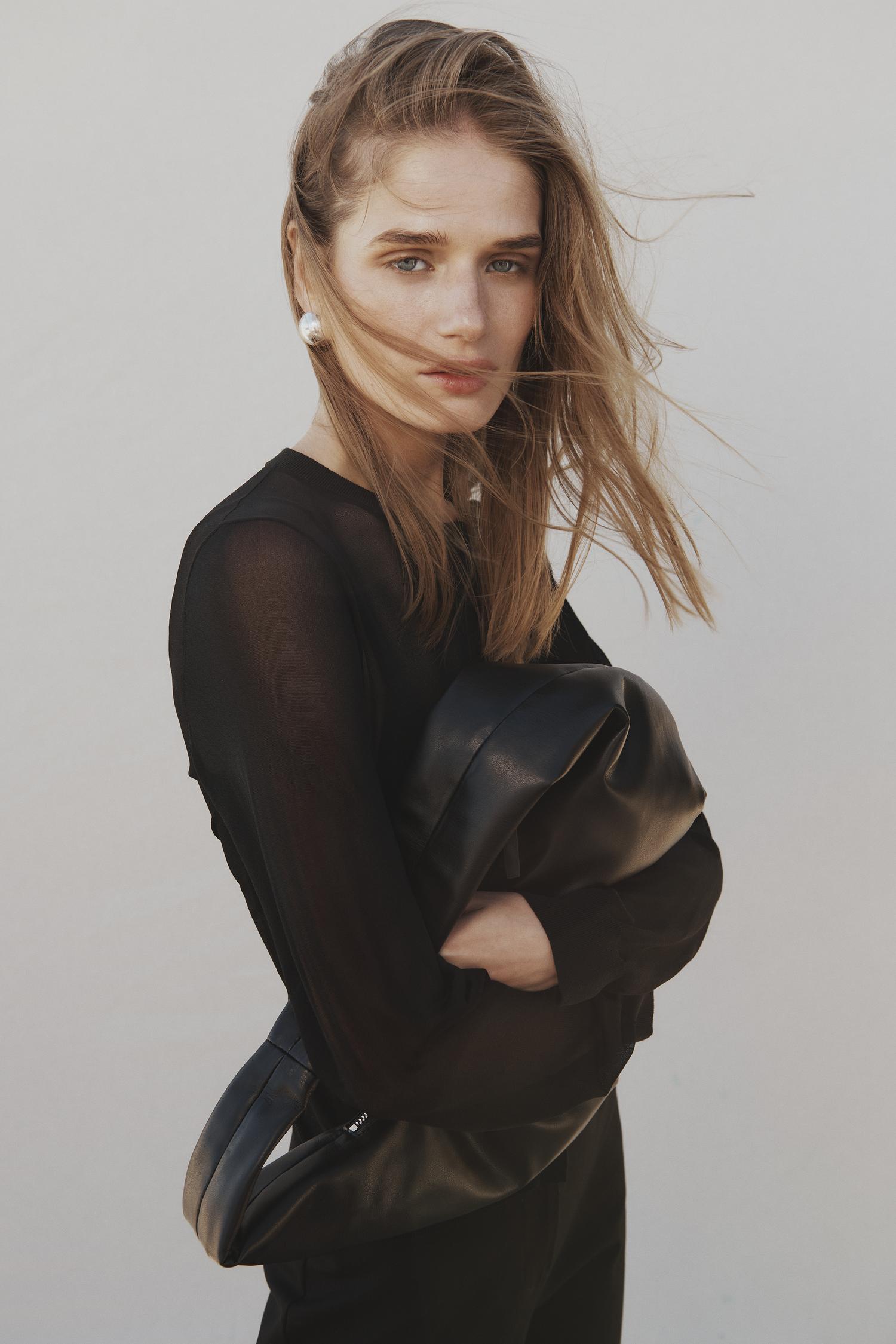 Blonde woman in black sheer top holding a black leather bag against a neutral backdrop in a fashion style portrait.