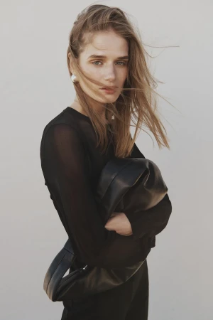 Blonde woman in black sheer top holding a black leather bag against a neutral backdrop in a fashion style portrait.