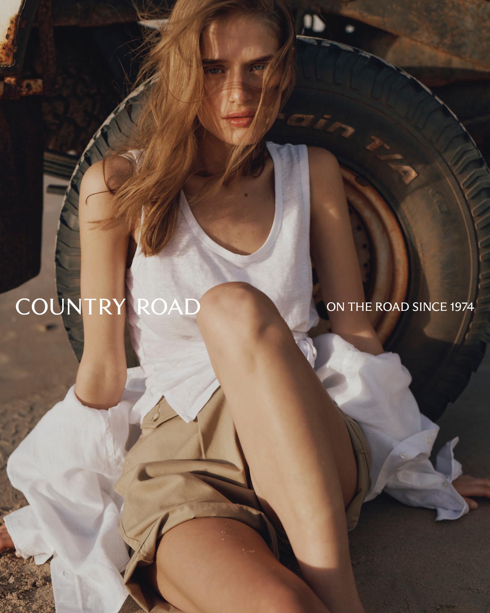 Fashion shot of a woman in a white tank top and khaki shorts sitting in sand near a black tire and an old vehicle on a sunny day.