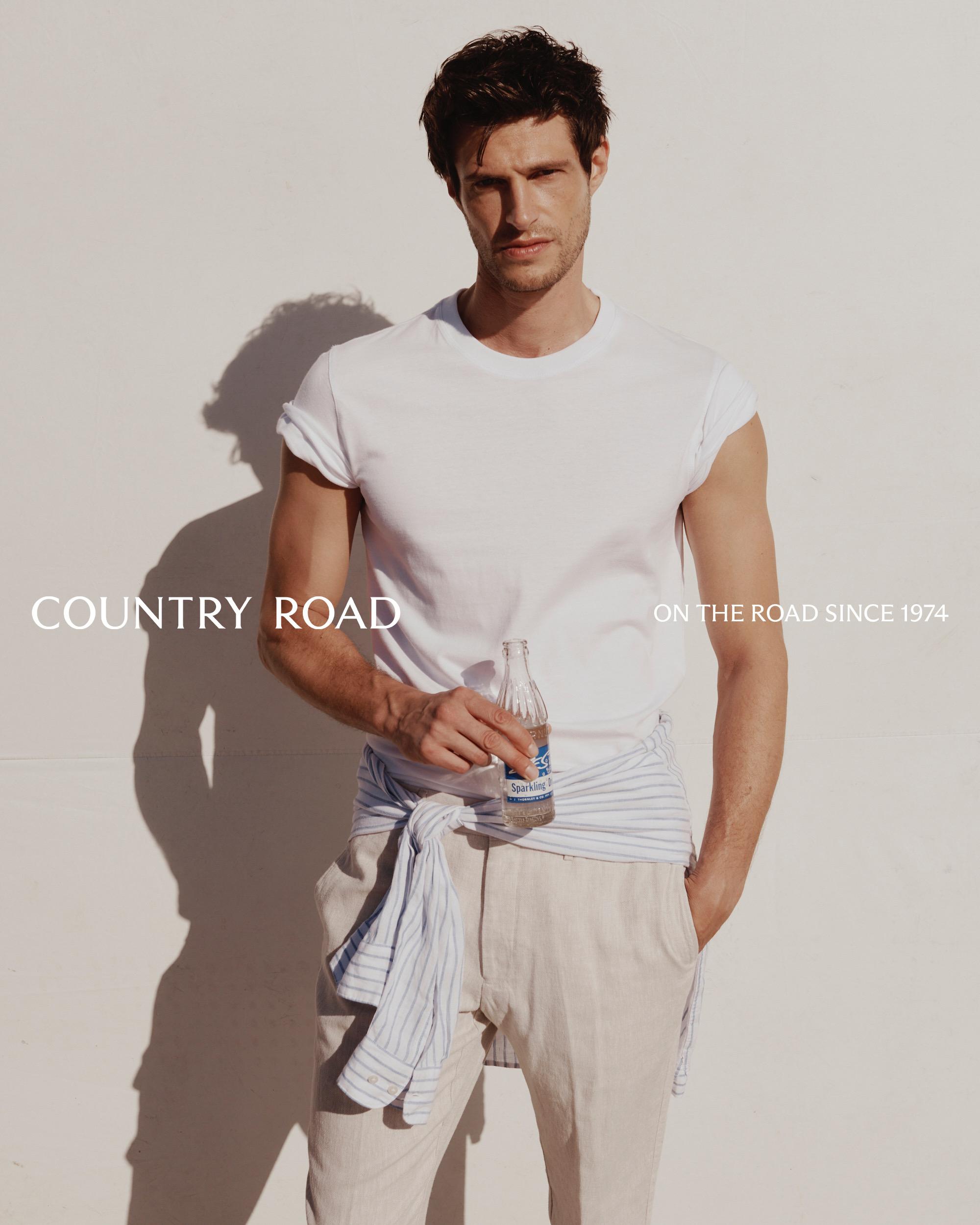 A man poses against a white wall, wearing a white T-shirt and pants, accessorized with a striped shirt tied around his waist.