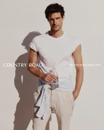A man poses against a white wall, wearing a white T-shirt and pants, accessorized with a striped shirt tied around his waist.