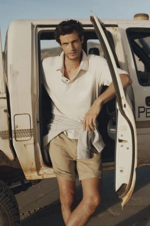 An adult male stands next to a white pickup truck wearing a polo shirt and khaki shorts, with a light blue sky overhead.