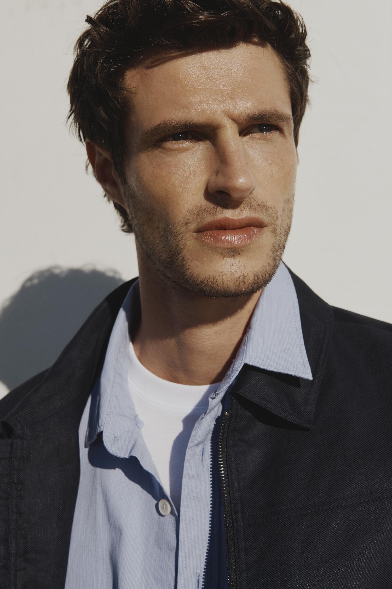 Portrait of a handsome adult male model in a blue collared shirt and black jacket against a white background in natural light.